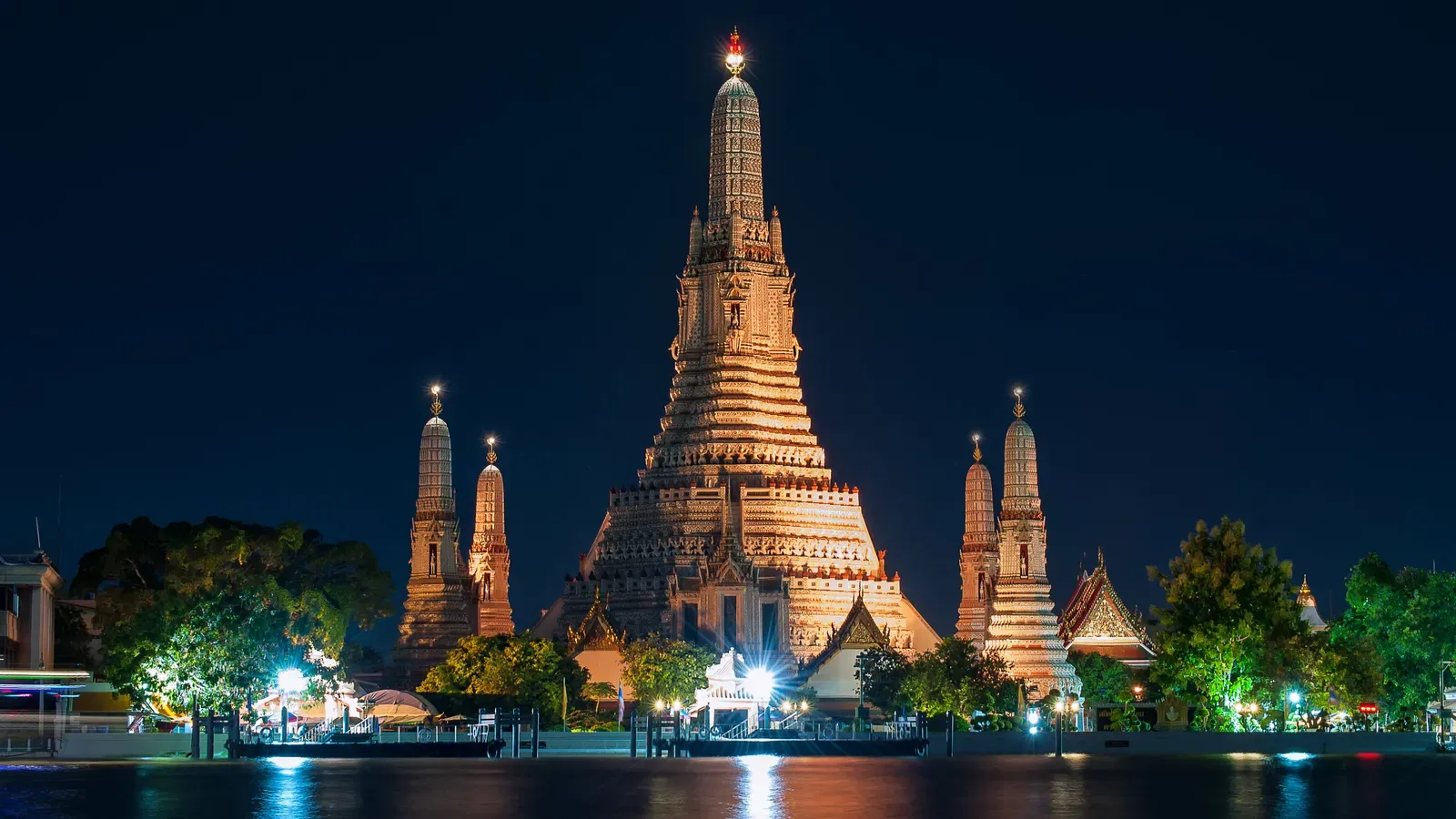 The floodlit central prang of Wat Arun and four smaller prangs rising above riverside trees, reflected in the Chao Phraya River at night