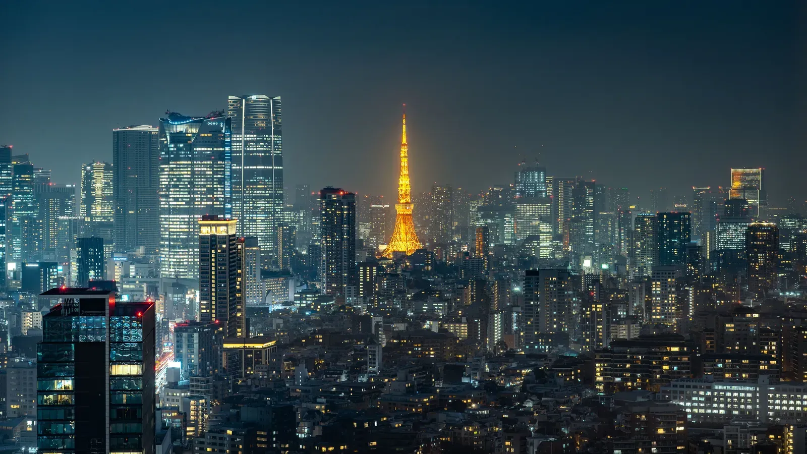 Tokyo Tower lit orange among the illuminated skyscrapers of Roppongi and Azabudai at night, above a dense spread of city lights