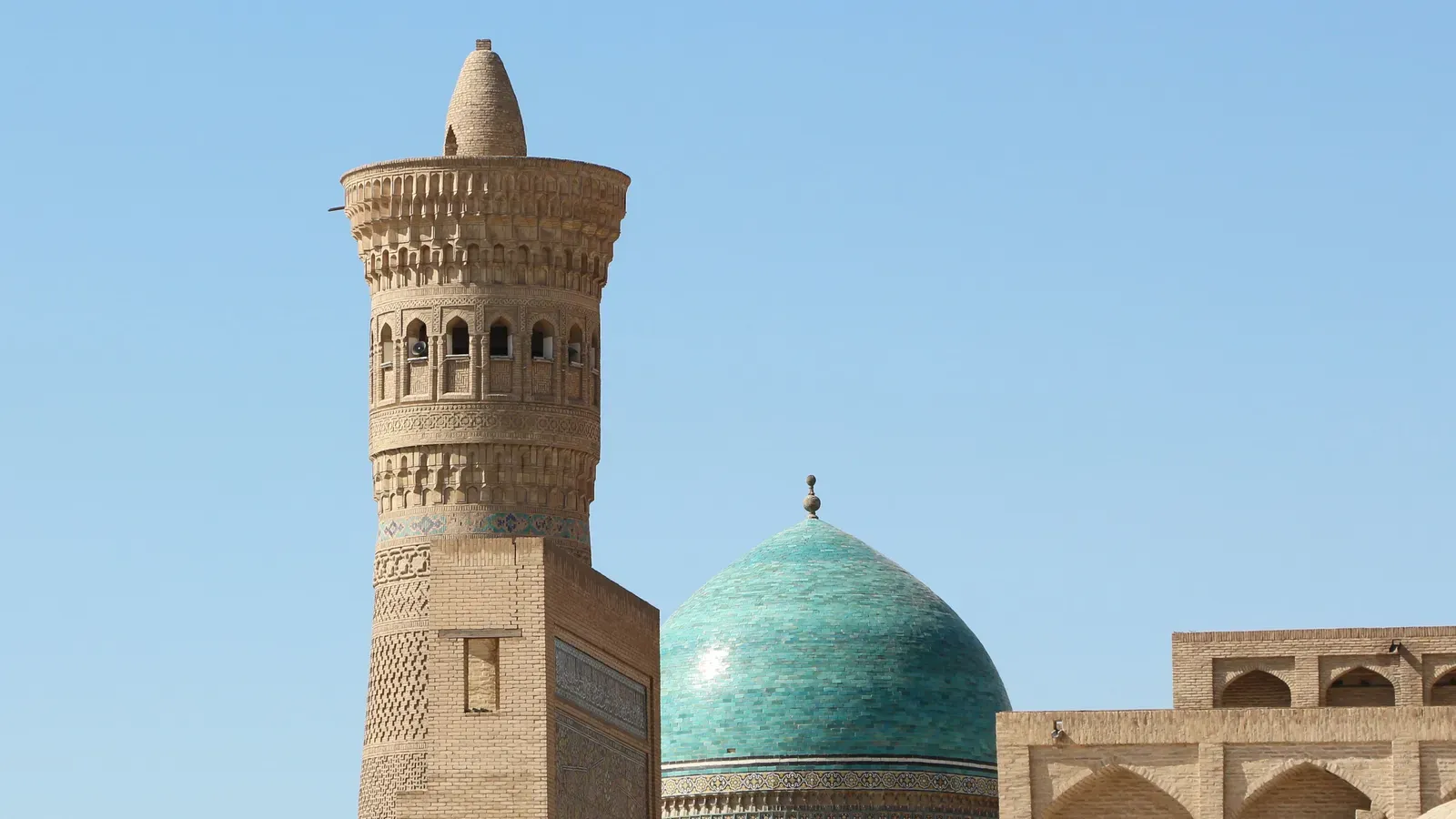The carved brick Kalyan Minaret beside the turquoise tiled dome of the Mir-i-Arab Madrasa under a clear blue sky in Bukhara