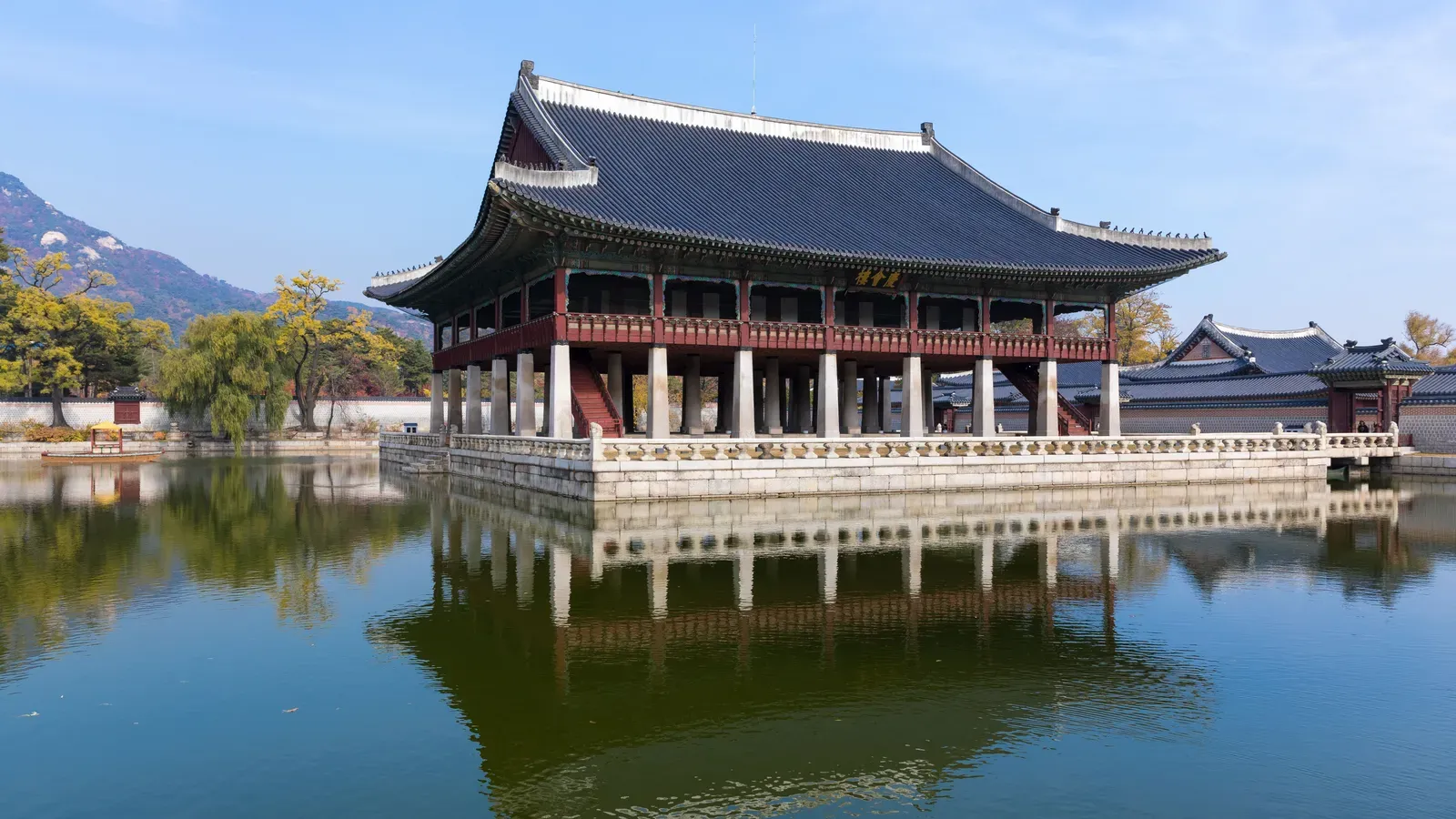 The two-storey Gyeonghoeru banquet pavilion on stone pillars reflected in its pond, with autumn trees and a mountain behind, at Gyeongbokgung Palace