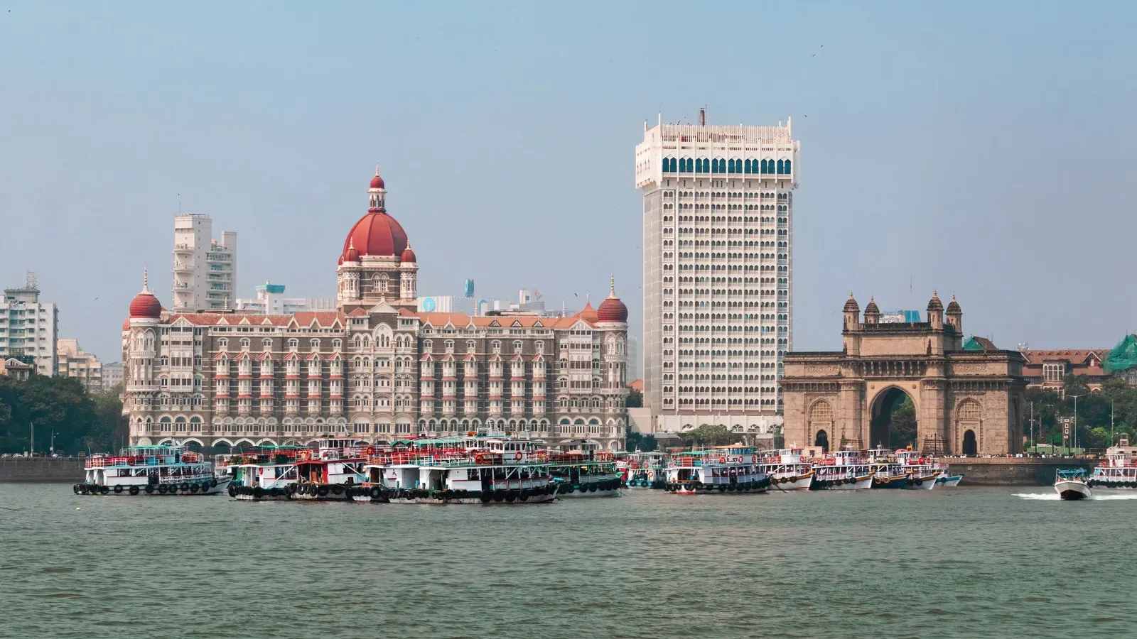 The Gateway of India arch beside the red-domed Taj Mahal Palace and the white Taj Tower, seen across Mumbai Harbour with passenger ferries moored in front