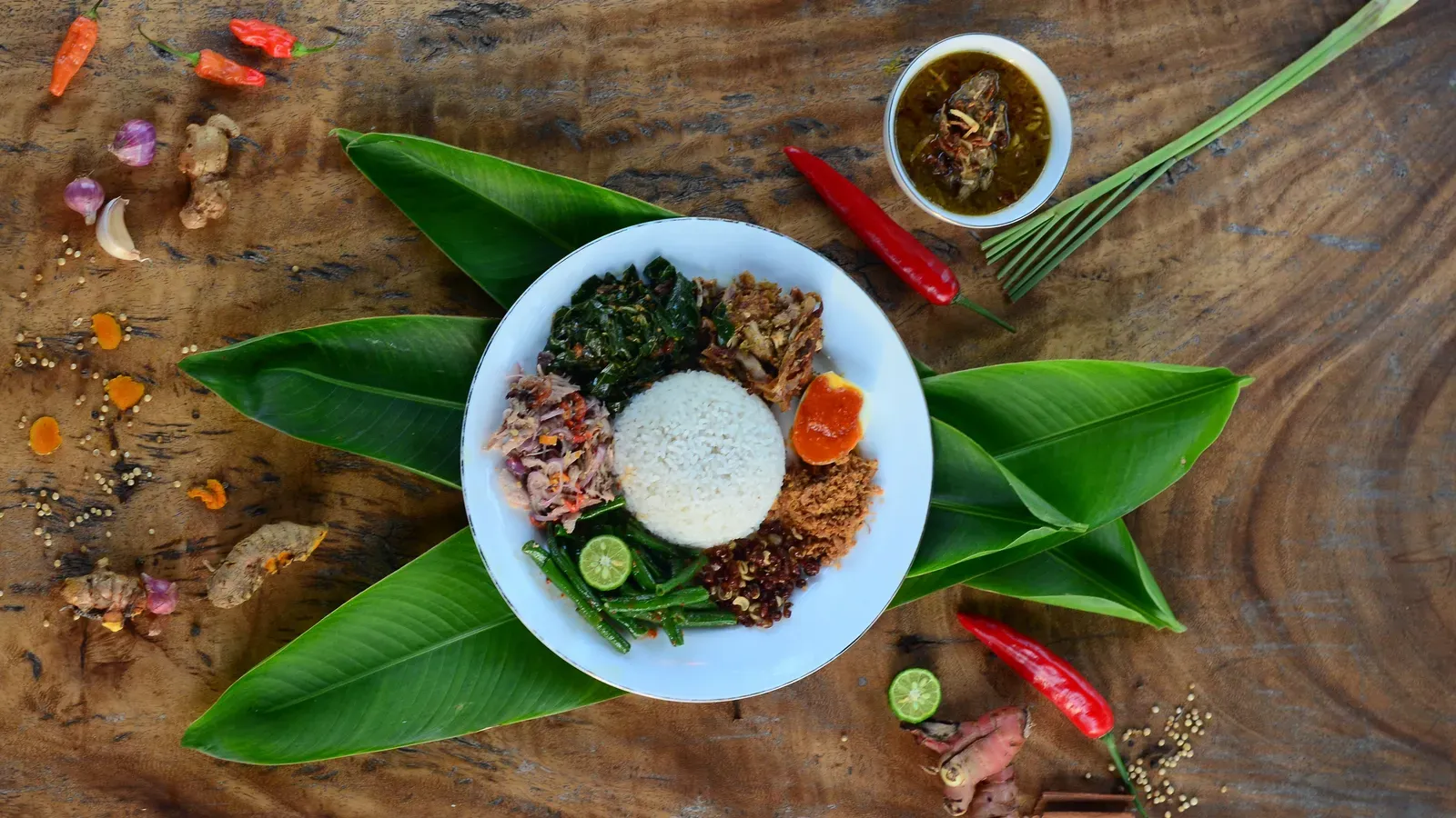 A plate of Balinese nasi campur with steamed rice, shredded chicken, long beans, greens and a sambal-topped egg, set on banana leaves on a wooden table with chillies, ginger and a bowl of chicken soup