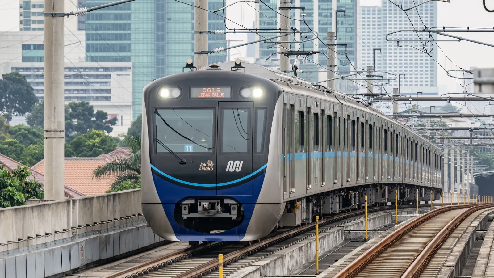 A blue and silver MRT Jakarta train approaching on the elevated track, with overhead wires, trees, tiled roofs and office towers behind
