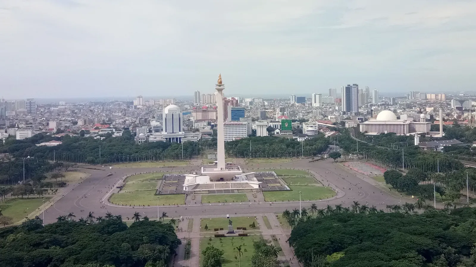 The National Monument, a tall white obelisk topped with a gold flame, standing in the open lawns of Merdeka Square, with Istiqlal Mosque's white dome and the Central Jakarta skyline behind