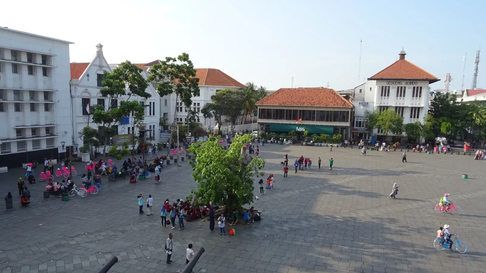 Fatahillah Square in Kota Tua seen from above, with visitors, colourful rental bicycles and a shade tree on the paved square, and Dutch colonial buildings including Cafe Batavia behind