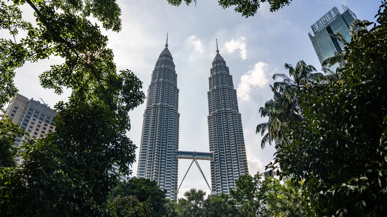 The Petronas Twin Towers and their skybridge rising above the trees and palms of KLCC Park under a partly cloudy sky