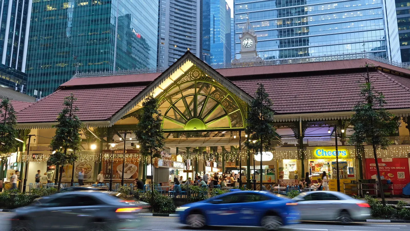 The Victorian cast-iron market hall of Lau Pa Sat lit up at dusk, with diners at tables under the eaves, office towers behind and cars blurred in the foreground