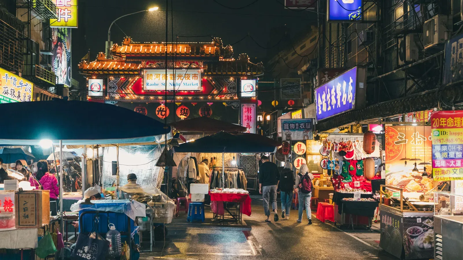 A wet street at night lined with food and clothing stalls under umbrellas, neon signs and red lanterns, with the lit gate of Raohe Street Night Market ahead